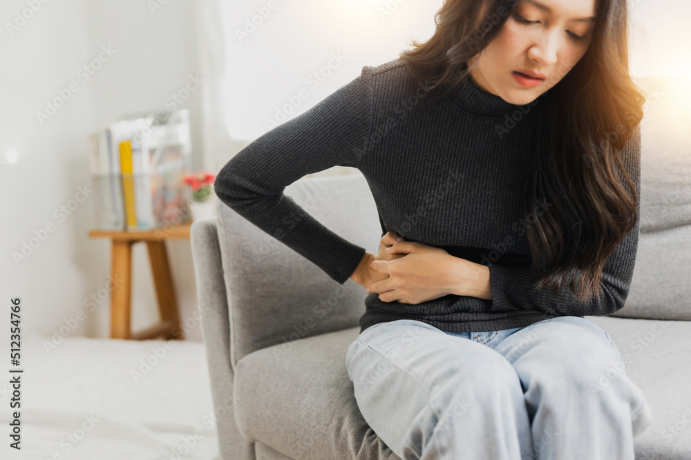 Naklejka premium Young woman suffering from strong abdominal pain while sitting on sofa at home