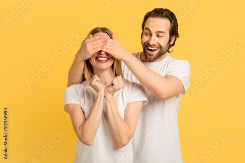 Cheerful glad millennial caucasian guy in white t-shirt closes eyes to woman for surprise