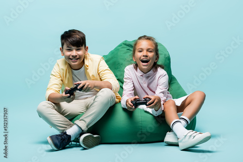 Quadro su tela KYIV, UKRAINE - JULY 2, 2021: Happy multiethnic preteen kids playing video game on beanbag chair on blue background