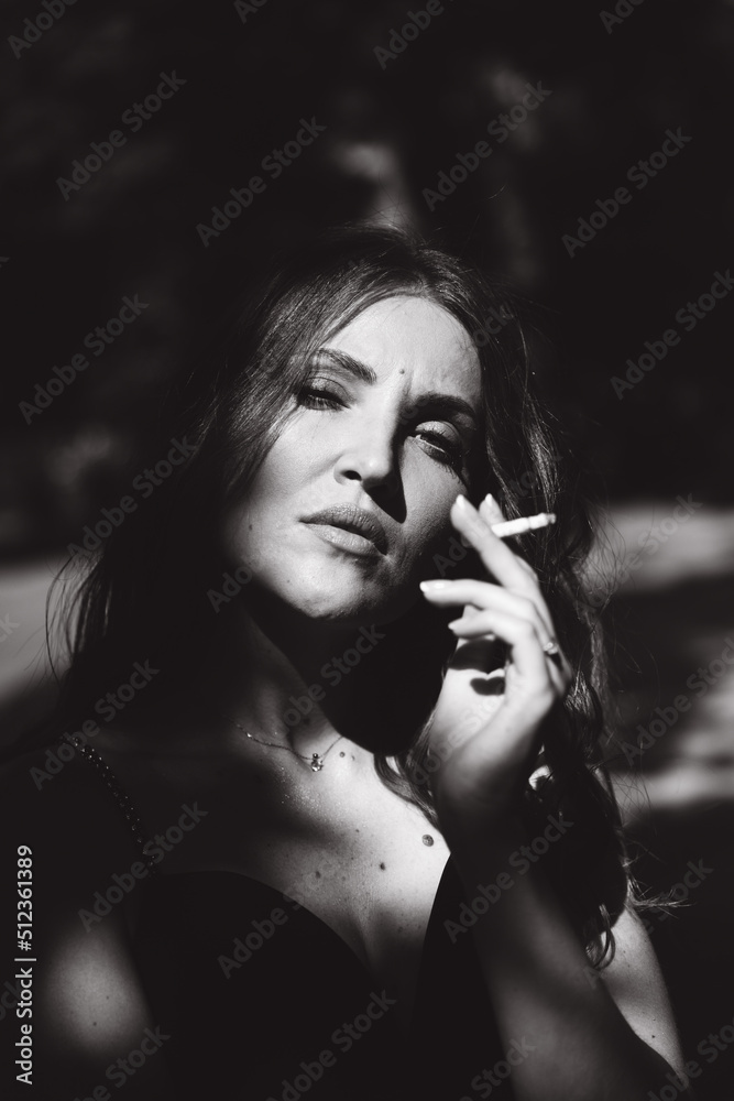 Fototapeta premium A beautiful girl with a hairstyle in a black dress on the street with a cigarette in her hands smokes, black and white photo.