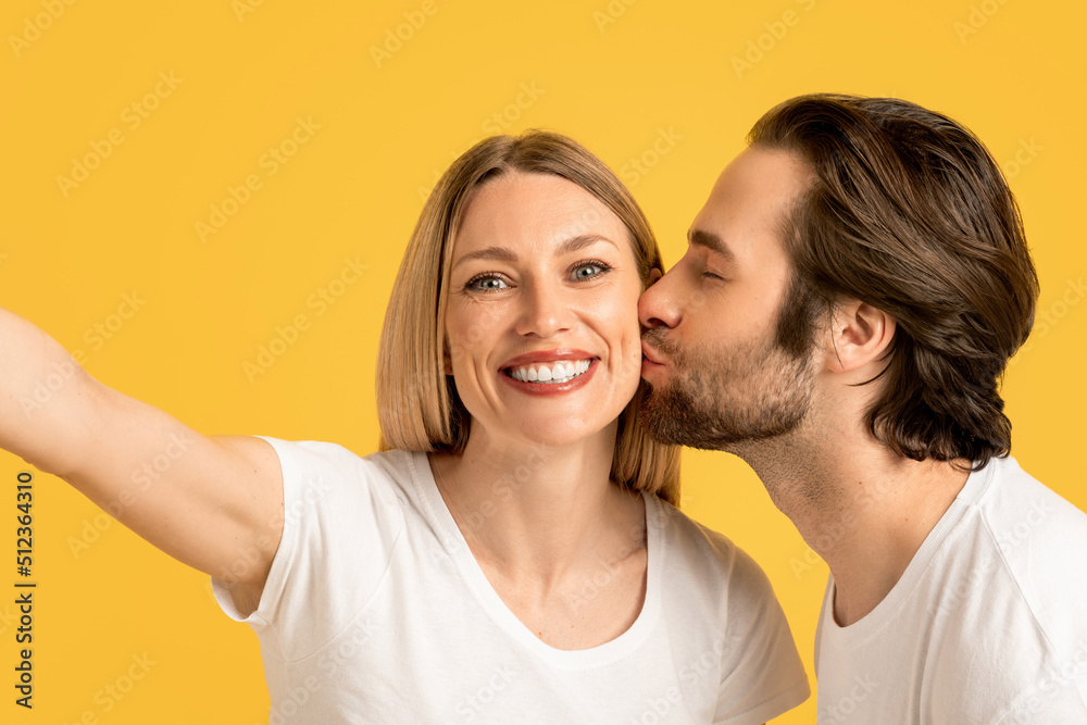 Happy millennial caucasian husband kissing wife on cheek in white t-shirt, lady make selfie Happy millennial caucasian husband kissing wife on cheek in white t-shirt, lady make selfie