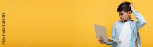 Pensive asian pupil with backpack looking at laptop isolated on yellow, banner.
