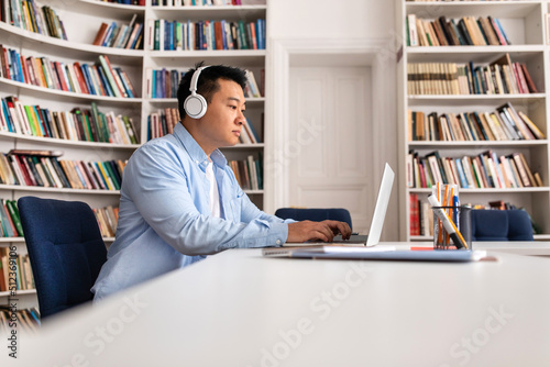 Asian Man Typing On Laptop Wearing Headphones Sitting In Library