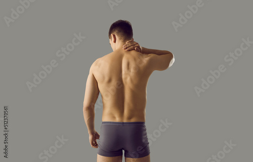 Rear view of naked sporty man in underwear isolated on grey studio background suffer from neck ache. Pain in upper back. Sexy toned guy in briefs struggle with backache or muscular spasm.