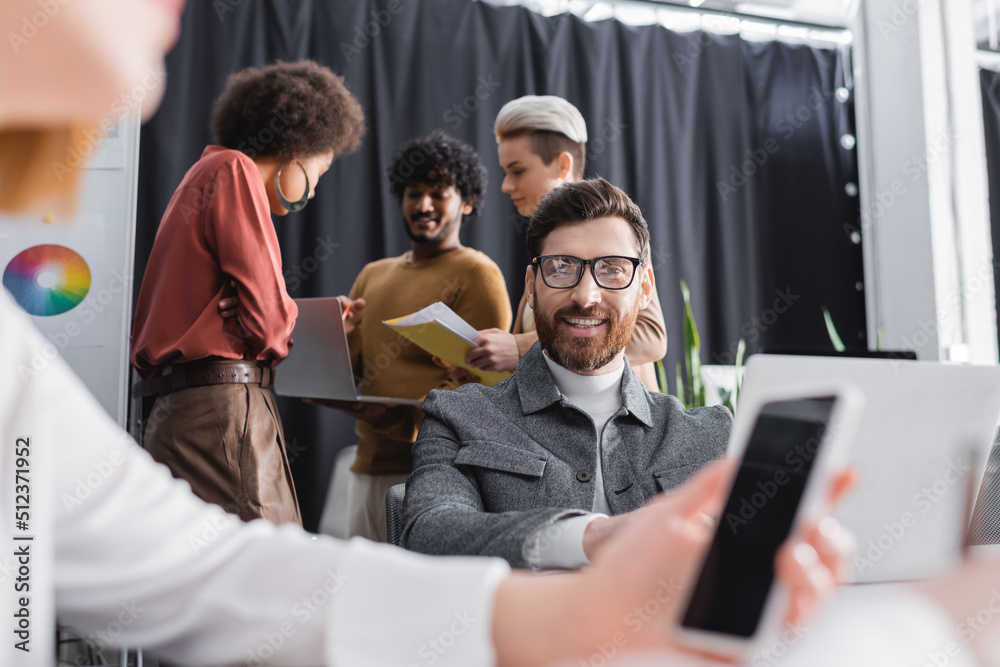 Obraz premium advertising manager in eyeglasses smiling near blurred colleague with smartphone.