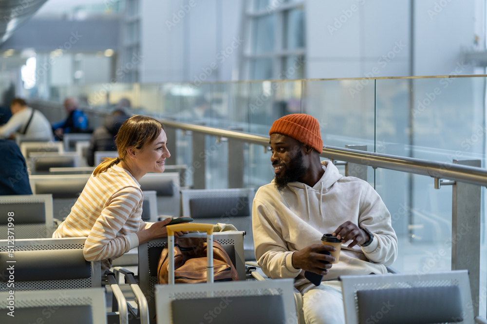 New acquaintances during flight delay. Outgoing African American hipster male with coffee and pretty Caucasian woman traveler talk about different topics sitting on chairs at terminal. Travel concept