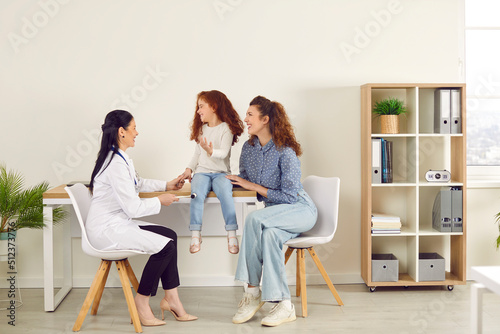 Friendly female doctor examining happy little patient in modern medical office interior. Cute cheerful preschool child and her mom laugh when professional neurologist uses hammer to test knee reflex photo
