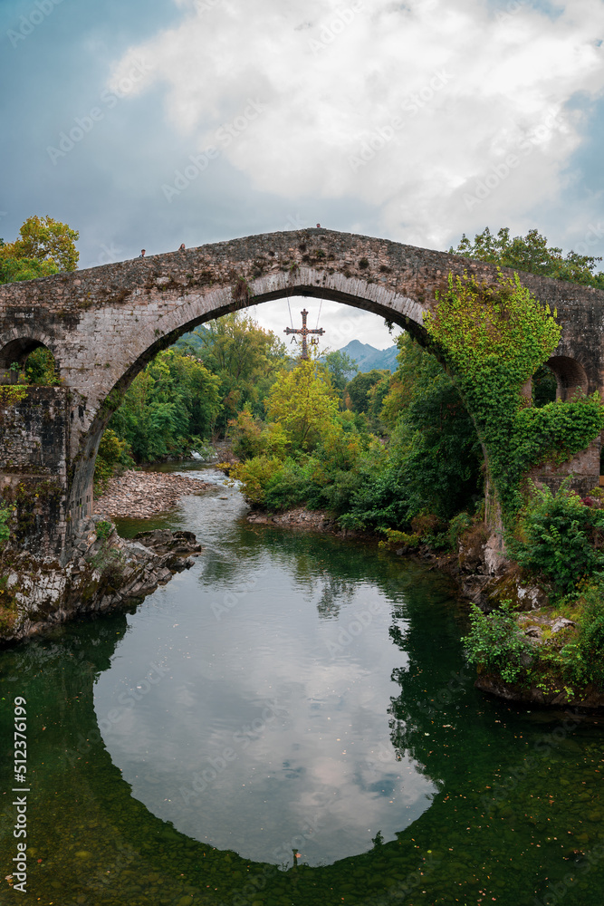 Famous Roman bridge over a lake, in Cangas de Onís, Asturias, with a lot of vegetation and a cross hanging over it. 