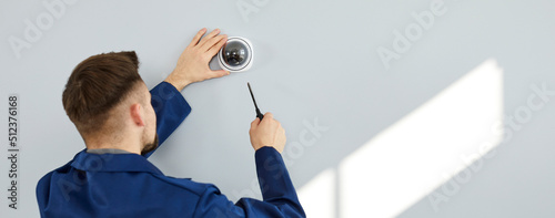 Young man installing modern security camera for theft deterrence inside new residential building. Repair service guy uses screwdriver to fit screws and adjust wall mounted CCTV surveillance dome cam photo