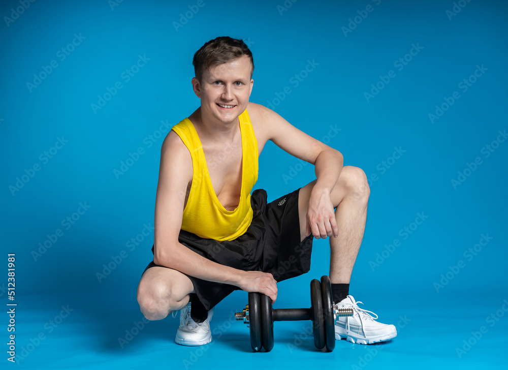 Obraz premium thin athlete in a yellow t-shirt and black shorts on a blue background. funny shakes muscles, goes in for sports, fitness