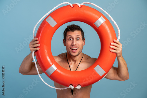 Male beach rescuer. Surprised bearded man holding inflated lifebuoy with wide opened mouth and being ready to rescue you isolated over blue background
