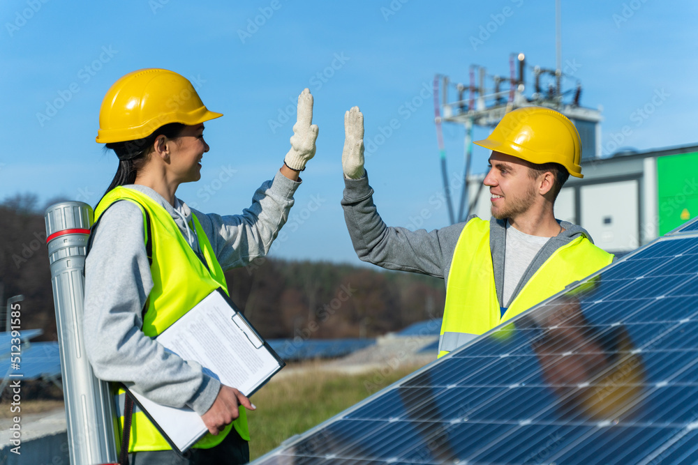 Two experienced solar panel workers in hardhat and vests giving five to each other and feeling happy while agreed about something
