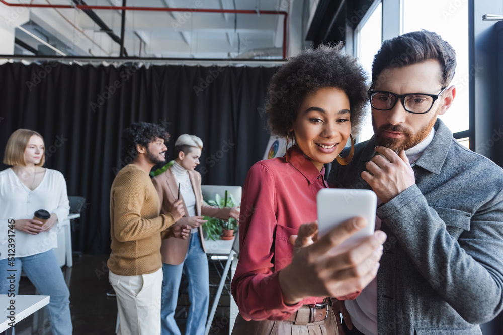 Fototapeta premium smiling african american woman showing smartphone to thoughtful colleague in ad agency.