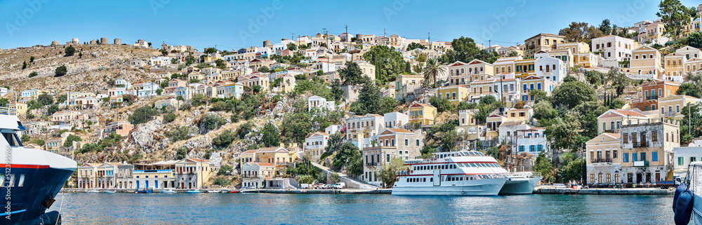 Multi-colored houses scattered on hills of Symi island view from water. Coastal town on turquoise sea shore in Greece. Tourist liners moor by seafront