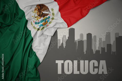 abstract silhouette of the city with text Toluca near waving colorful national flag of mexico on a gray background. photo