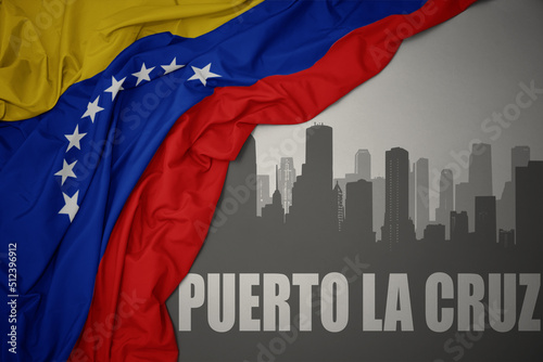 abstract silhouette of the city with text Puerto la Cruz near waving colorful national flag of venezuela on a gray background.