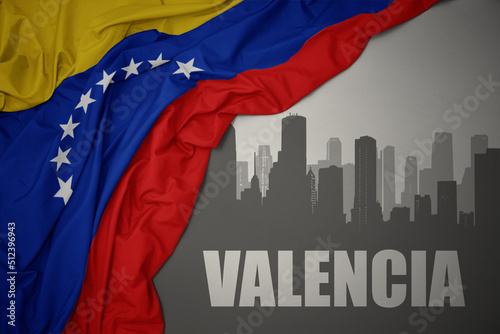 abstract silhouette of the city with text Valencia near waving colorful national flag of venezuela on a gray background.