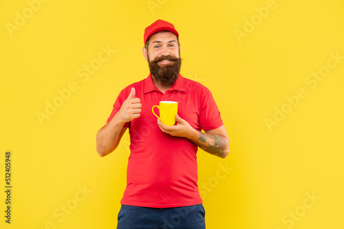 Happy bearded man in casual red cap and tshirt giving thumb holding mug yellow background, tea