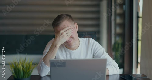 Upset young man covering face with palm in despair after reading bad news on laptop photo