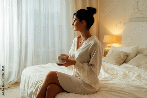 Lady enjoy good morning, weekend and vacation, delicious coffee at free time after great sleep. Satisfied pretty caucasian woman sits on white bed in bedroom