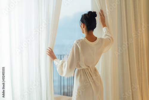 Morning of beautiful young woman opening curtains in bedroom and enjoying of the amazing views. Back view of the young lady