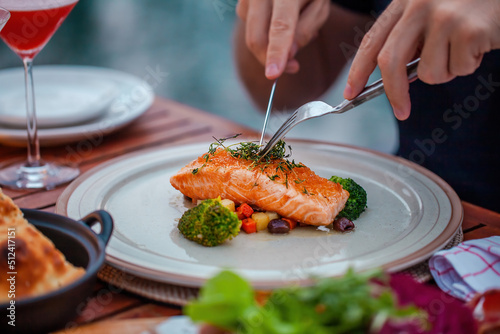 Man on luxury dinner served on table with fresh seafood, cold cuts and cocktail glass in luxury outdoor restaurant. Man hands cutting grilled salmon. Healthy food