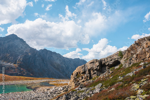 Lovely autumn view from rocks to turquoise mountain lake against high rocky mountain top in sunlight under clouds in blue sky. Sunlit beautiful small lake and large mountain range. Vivid autumn colors