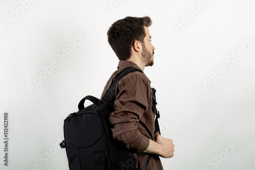 Young man wearing brown shirt posing isolated over white background student wearing a backpack with headphones around his neck. Back view photo.