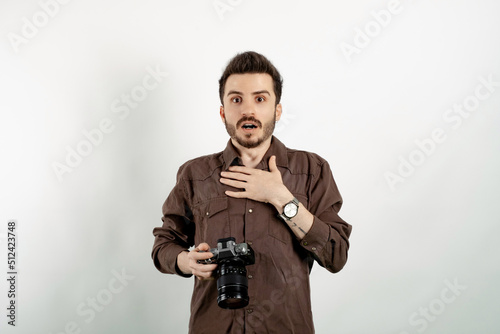 Handsome young man wearing casual clothes posing isolated over white background feeling shocked and surprised, smiling, taking hand to heart while holding dslr camera.