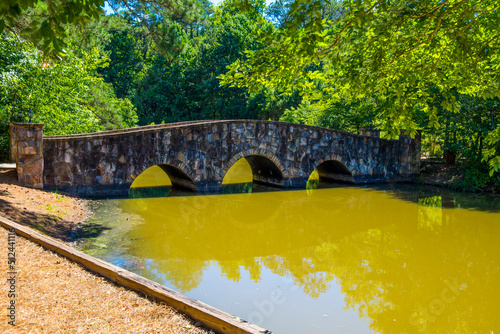 a gorgeous summer landscape at the pond with a stone bridge over the silky brown water surrounded by lush green trees, grass and plants with blue sky and clouds at Mill Creek Pond in Alpharetta photo