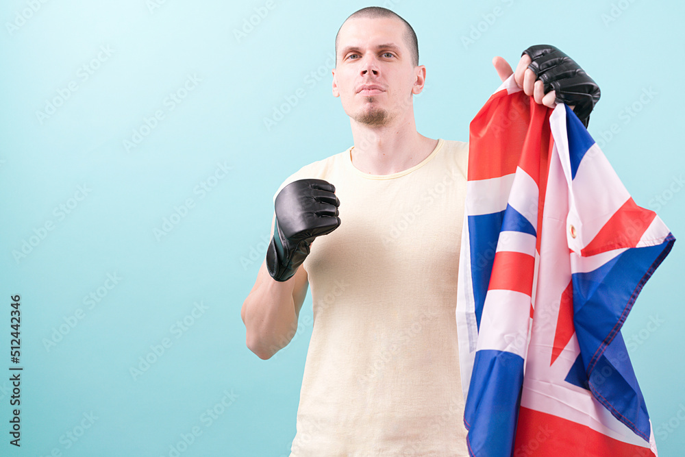 A confident MMA fighter in black gloves holds a UK flag ready to fight on a blue background. Fighting stance. Pose. Knockout. Bald. Mixed Martial Arts. Hit. Heavy. Tough. Hitting. Win. Goal