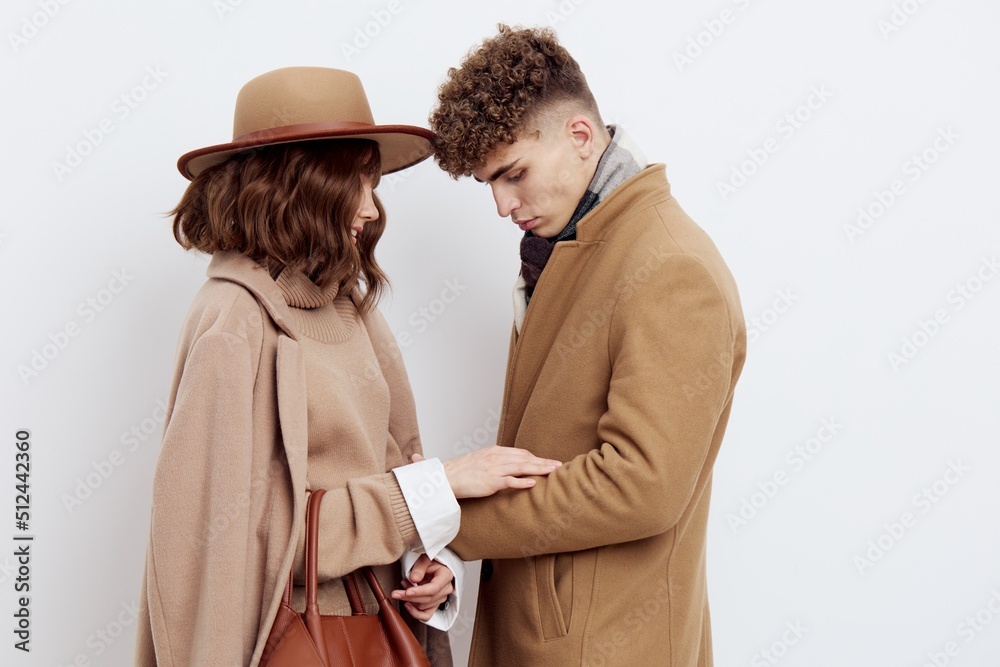 a cute couple stands on a light background dressed in coats and sweaters, a man stands against a woman and hugs her in front a cute couple stands on a light background dressed in coats and sweaters, a man stands against a woman and hugs her in front