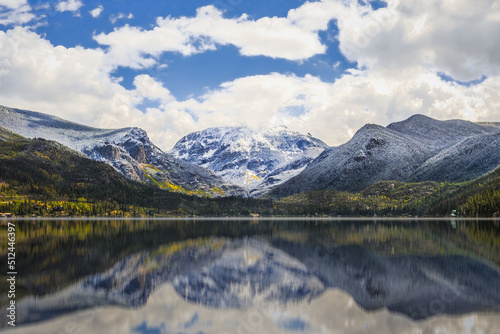 Picturesque view of Colorado mountain range covered with light snow on top reflecting in calm water; blue sky with clouds in background