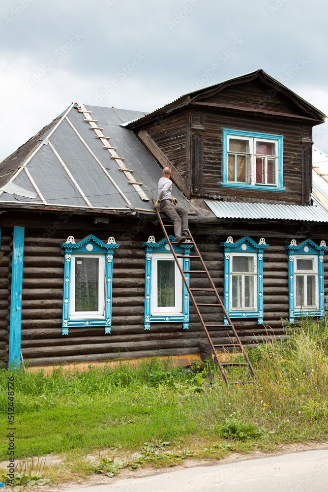 Middle-aged man sitting back on metal ladder repairing roof of old wooden brown house carved windows