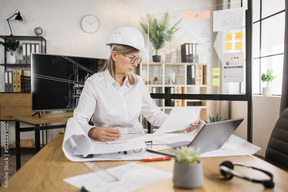 Senior woman builder, engineer in white shirt and hard hat on desk in office on background of large TV screen with, working on laptop while looking on papers or sketches. Senior woman builder, engineer in white shirt and hard hat on desk in office on background of large TV screen with, working on laptop while looking on papers or sketches.
