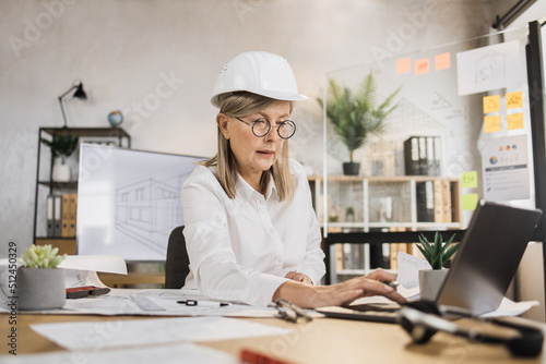 Portrait of professional caucasian mature female architect using laptop computer, working on construction plan, using sketches at her workplace on light modern office.