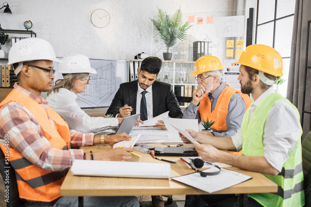 Multiracial men and women in formal outfit and helmets sitting at table with lots of blueprints during important strategic session. Business people working on common construction project. Multiracial men and women in formal outfit and helmets sitting at table with lots of blueprints during important strategic session. Business people working on common construction project.