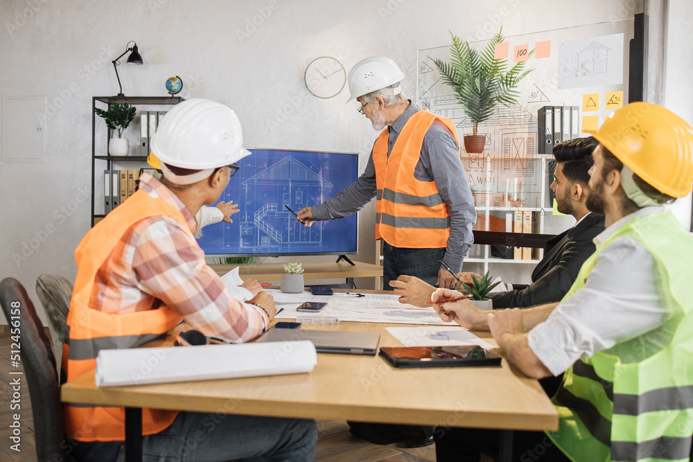 Senior bearded man in uniform and helmet presenting new construction project to colleagues on tv screen at office. Three men and one woman sitting at table and listening their competent partner. Senior bearded man in uniform and helmet presenting new construction project to colleagues on tv screen at office. Three men and one woman sitting at table and listening their competent partner.