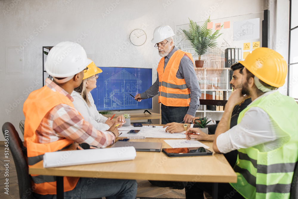 Senior bearded man in uniform and helmet presenting new construction project to colleagues on tv screen at office. Three men and one woman sitting at table and listening their competent partner. Senior bearded man in uniform and helmet presenting new construction project to colleagues on tv screen at office. Three men and one woman sitting at table and listening their competent partner.