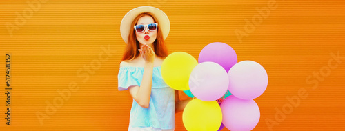 Summer portrait of happy woman blowing her lips with colorful balloons having fun wearing straw hat on orange background, blank copy space for advertising text © rohappy