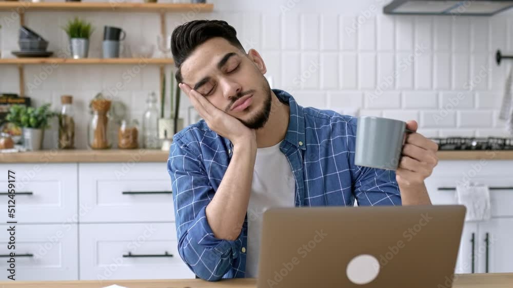 Bored indian or arabian guy, freelancer, designer, working remotely from home, tired of boring online work, suffering from chronic fatigue or overwork, holds a cup of coffee in his hand, falls asleep Bored indian or arabian guy, freelancer, designer, working remotely from home, tired of boring online work, suffering from chronic fatigue or overwork, holds a cup of coffee in his hand, falls asleep