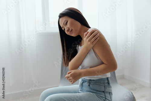 Obraz na płótnie Tormented from shoulder dislocation fractures pain after lifting weights in the wrong position tanned beautiful young Asian woman touching painful shoulder at home interior living room