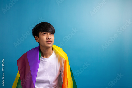 Asian young man with pride LGBT rainbow flag on shoulder against blue color background. A man with gay pride flag with copy space in studio.