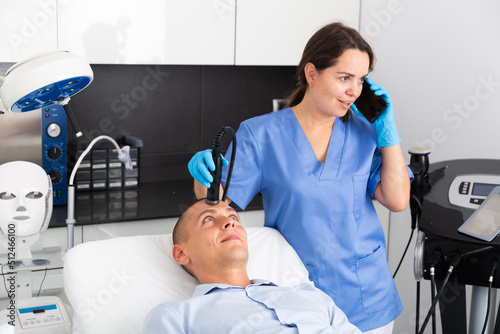 Cheerful female beautician talking on mobile phone while performing hardware facial treatment to male client in aesthetic medicine office. Unprofessional behavior concept photo