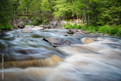 The blurred water of Duchesnay Falls flows down a river  under a stone footbridge through a forest in North Bay  Ontario during sunset.