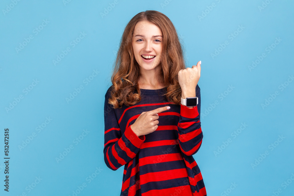 Fototapeta premium Look at clock, no rush! Portrait of positive woman wearing casual style sweater pointing wrist watch and expressing optimism about time, not busy. Indoor studio shot isolated on blue background.