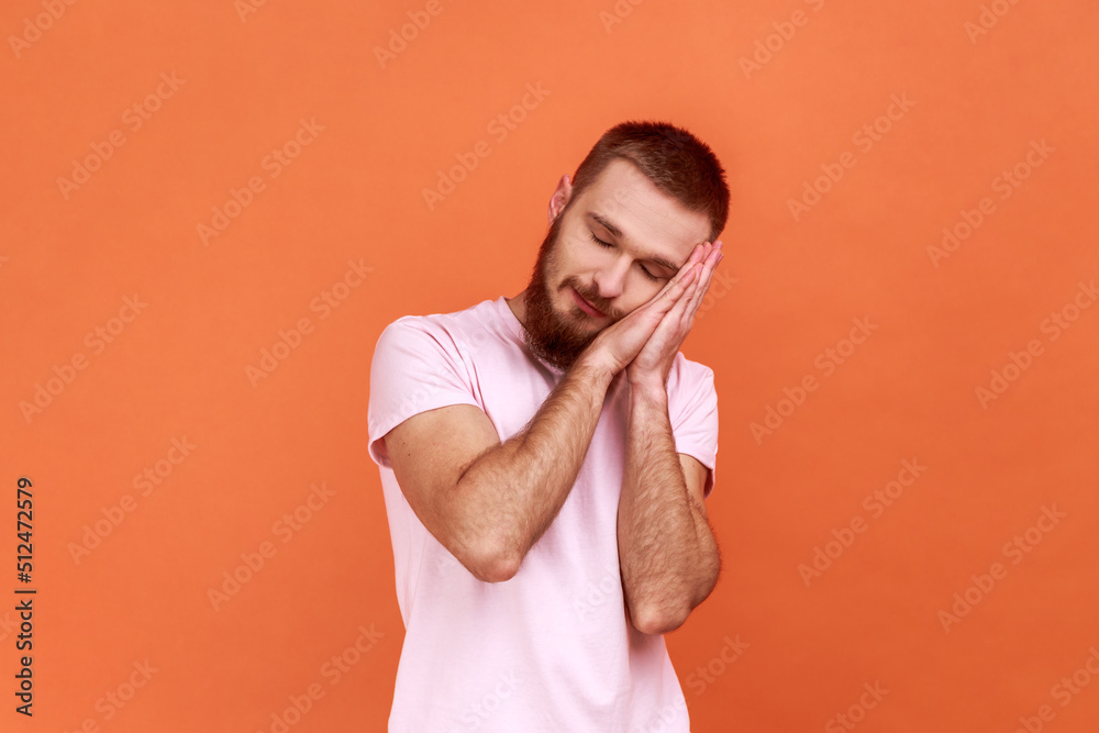 Portrait of bearded man sleeping laying down on her palms and smiling pleased, having comfortable nap and resting, dozing off, wearing pink T-shirt. Indoor studio shot isolated on orange background. Portrait of bearded man sleeping laying down on her palms and smiling pleased, having comfortable nap and resting, dozing off, wearing pink T-shirt. Indoor studio shot isolated on orange background.