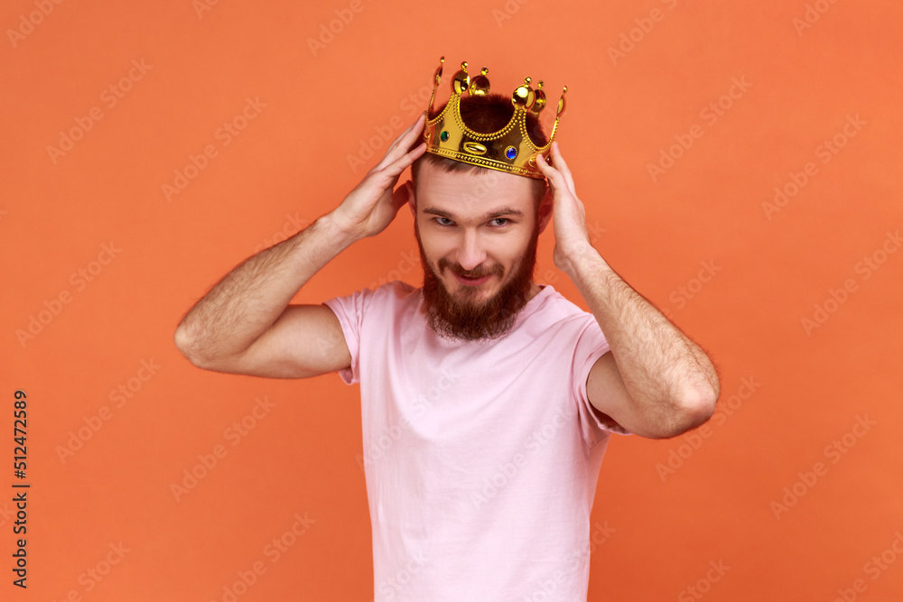 Portrait of handsome bearded man wearing golden crown, looking with positive facial expression, privileged status, wearing pink T-shirt. Indoor studio shot isolated on orange background. Portrait of handsome bearded man wearing golden crown, looking with positive facial expression, privileged status, wearing pink T-shirt. Indoor studio shot isolated on orange background.