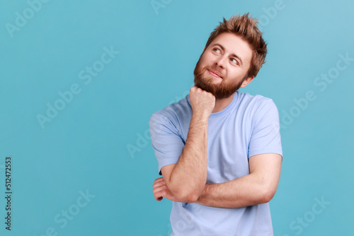 Portrait of smiling happy bearded man looking away with pensive expression, dreaming pleasant thoughts, fantasizing and making wish. Indoor studio shot isolated on blue background.