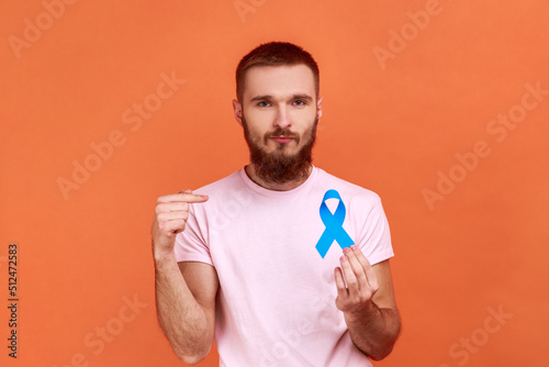 Portrait of serious bearded man pointing finger at blue awareness, disease symbol, looking at camera, support, wearing pink T-shirt. Indoor studio shot isolated on orange background. © khosrork